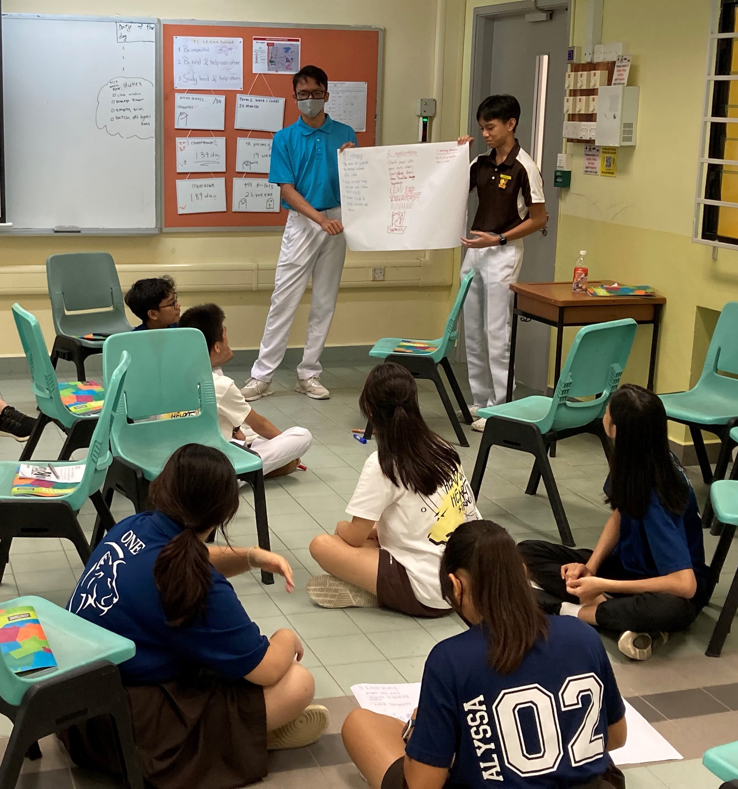 Two students hold a large poster while classmates sit on the floor in a classroom during a group presentation.